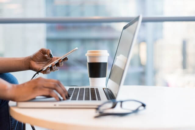 Young adult woman with phone in her hand and typing on the computer learning how to stop procrastinating at work