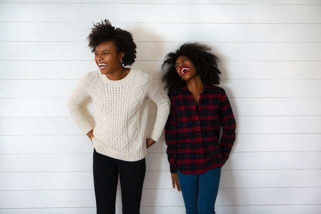 Two young adult Black female women are standing together and laughing. Demonstrating making friends with social anxiety.