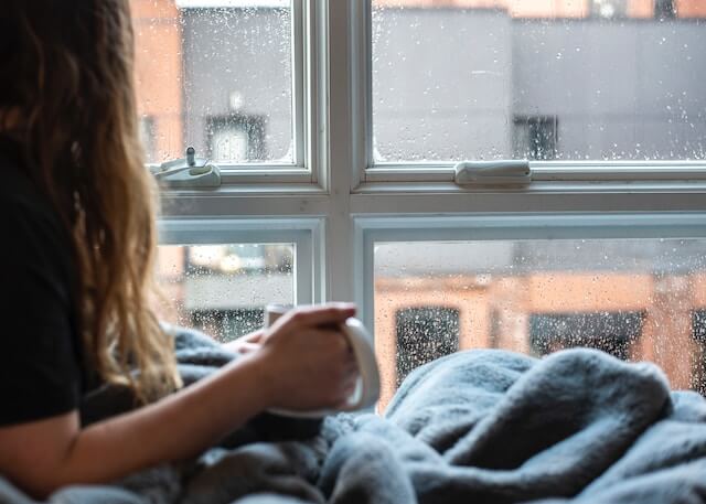 Young adult woman sitting by the window with a cup in her hand. She may be doing nervous habits and anxiety habit of staying in her comfort zone.